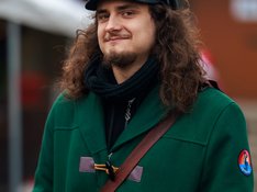 Portrait eines jungen Mannes mit langen braun gelockten Haaren, einer schwarzen Schiebermütze und grünem Mantel.